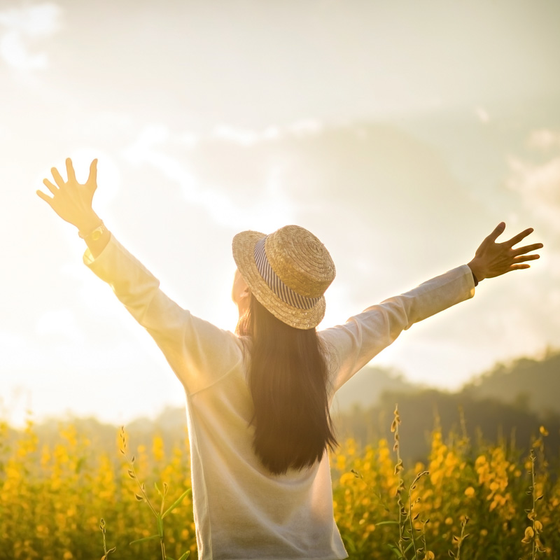 Photo of a woman from the back with outstretched arms facing the sun and a field of yellow flowers